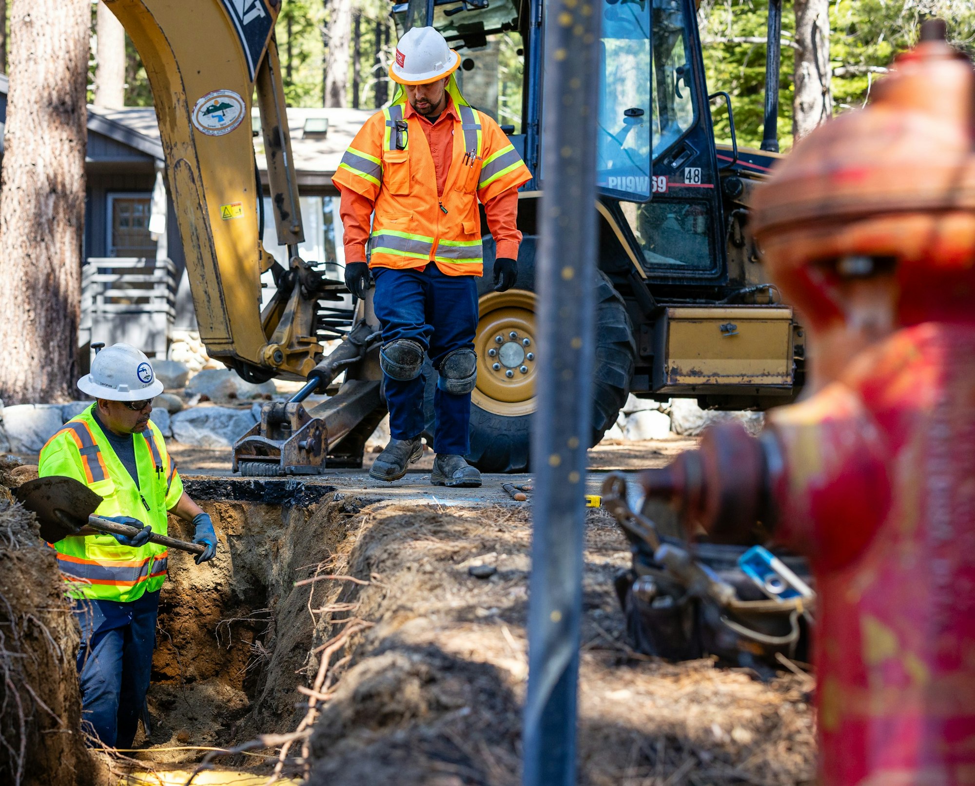 Two workers in high-vis near an excavator and a fire hydrant, engaged in construction or repair work.