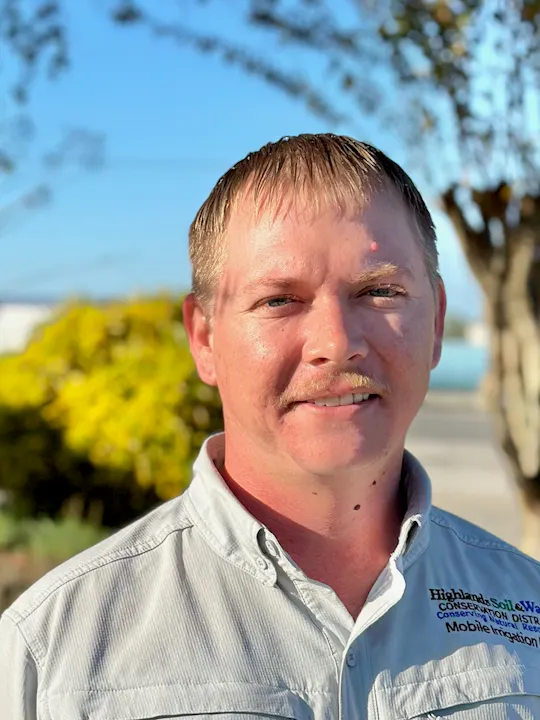 The image features a man with short hair and a mustache, smiling outdoors with greenery and a clear blue sky in the background.