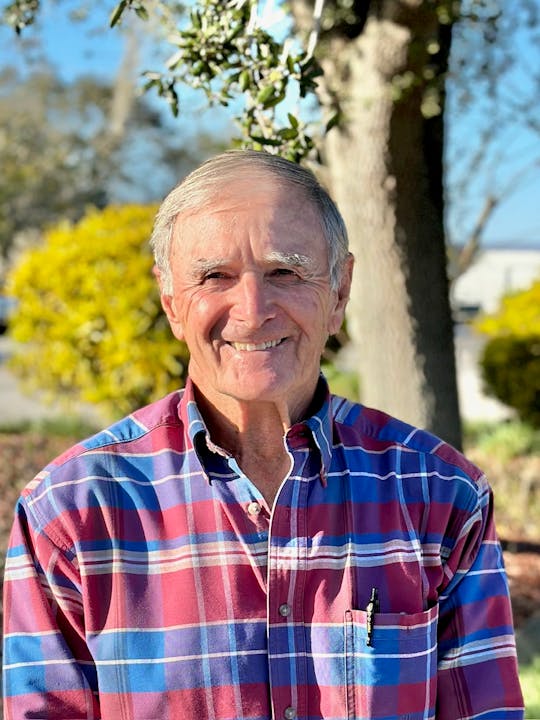 A smiling older man in a plaid shirt stands outdoors, surrounded by greenery and a clear blue sky.