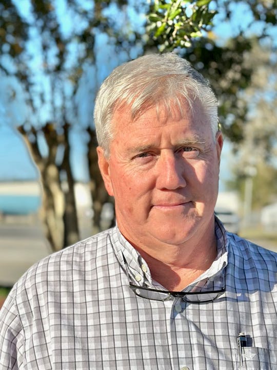 A man with gray hair is standing outdoors, wearing a checked shirt and holding glasses, with trees in the background.