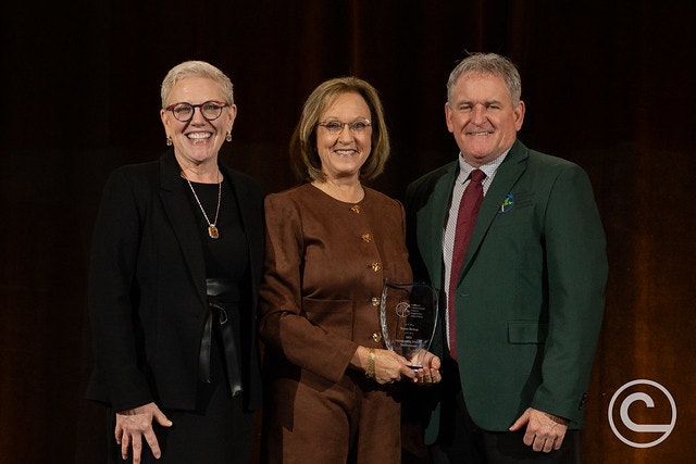 Three individuals stand together on stage, one holding an award, in a formal setting with a warm background.