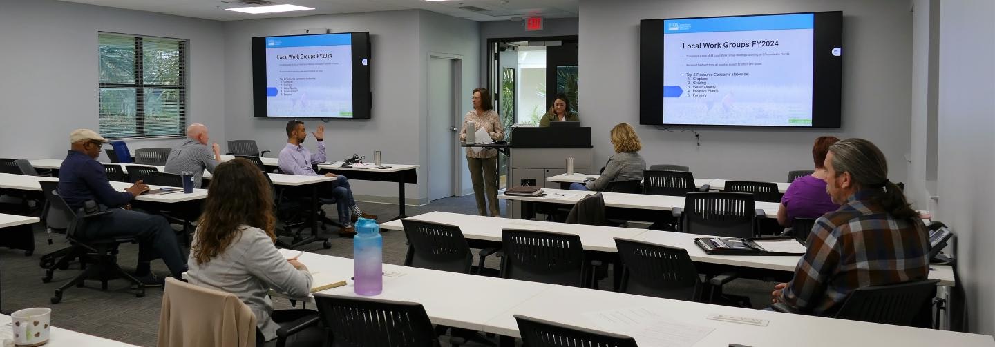 A professional meeting room with attendees seated, and two presenters standing near screens showing a presentation.