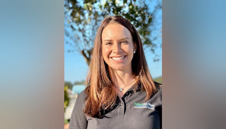 A smiling woman in a black polo with a conservation organization logo, set against a sunny outdoor background.