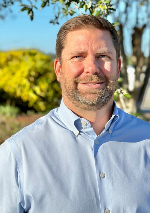 A man with a beard is smiling outdoors, wearing a light blue button-up shirt, with greenery in the background.