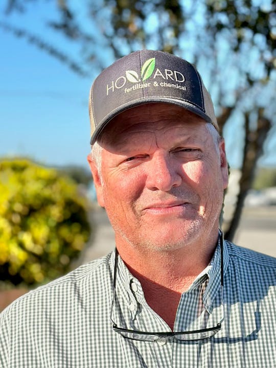 A man wearing a "Howard Fertilizer & Chemical" hat and a checkered shirt stands outdoors, smiling in front of greenery.