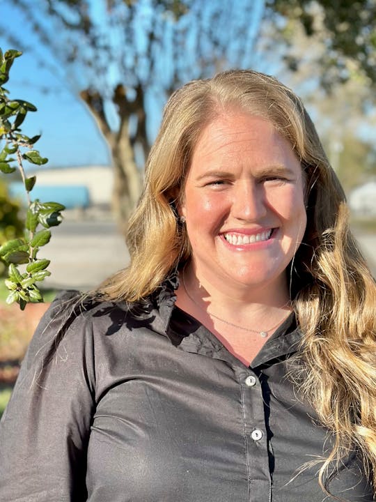 A smiling woman with long hair is outdoors, wearing a black shirt, with trees and a bright sky in the background.