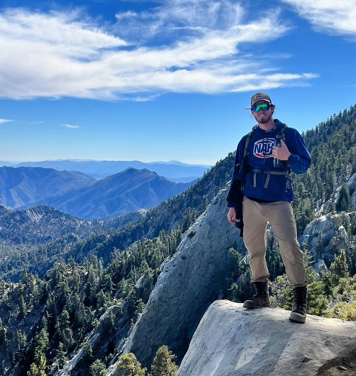 A person standing on a mountain ledge with a scenic view of mountains and trees in the background under a partly cloudy sky.