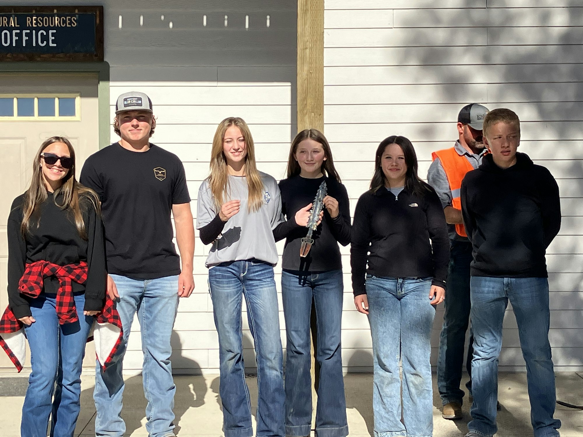 A group of six young people stands outside an office, wearing casual clothes and smiling, with one holding an object.
