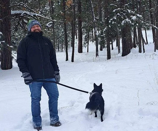 A man with a beanie and a dog on a leash standing in a snowy forest.