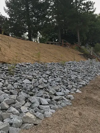 A slope with rocks at the base, trees at the top, and a dirt path alongside. It appears to be an erosion control measure.