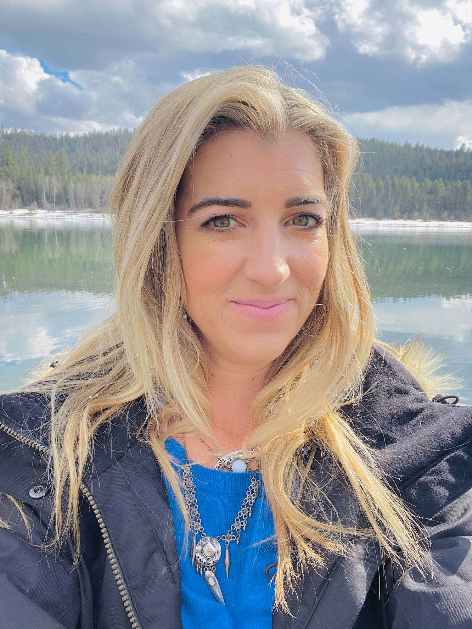 A person with a blue top and black jacket, smiling in front of a serene lake surrounded by forest and a cloudy sky.