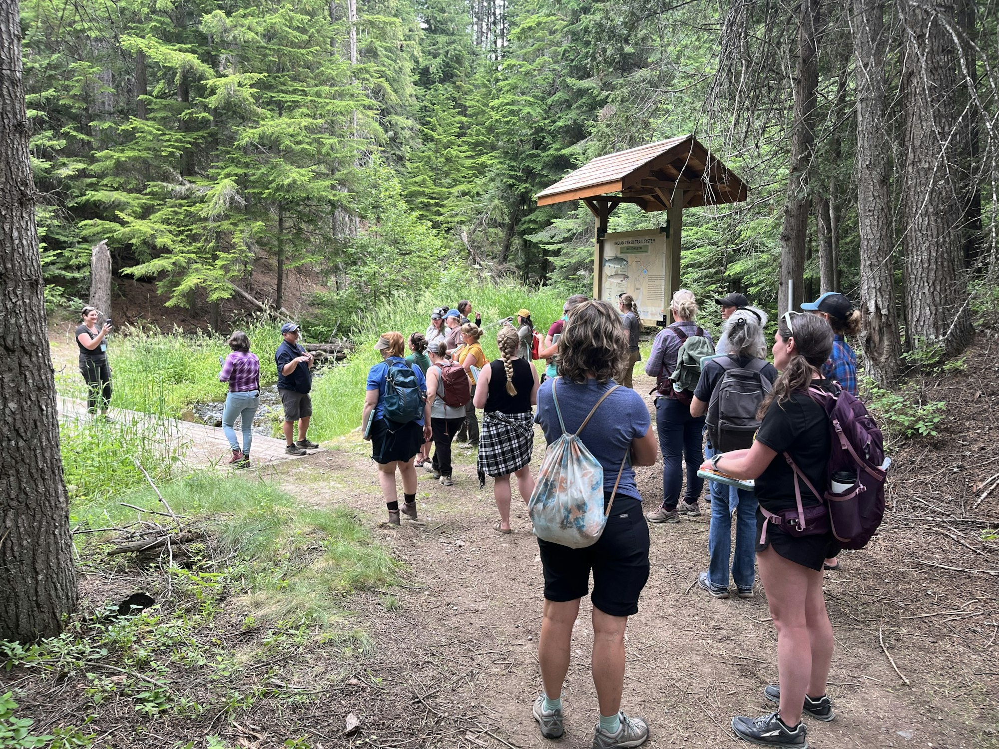 A group of people stands on a forest trail, listening to a person speaking, with trees and a wooden sign nearby.