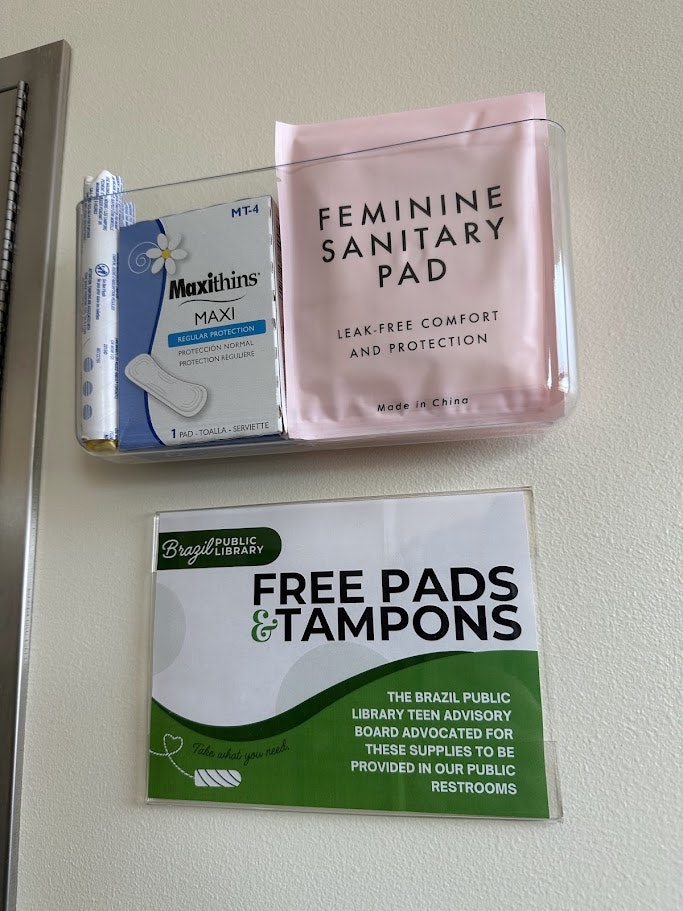 Sanitary pads in a dispenser with a sign offering free pads and tampons at the Brazil Public Library.
