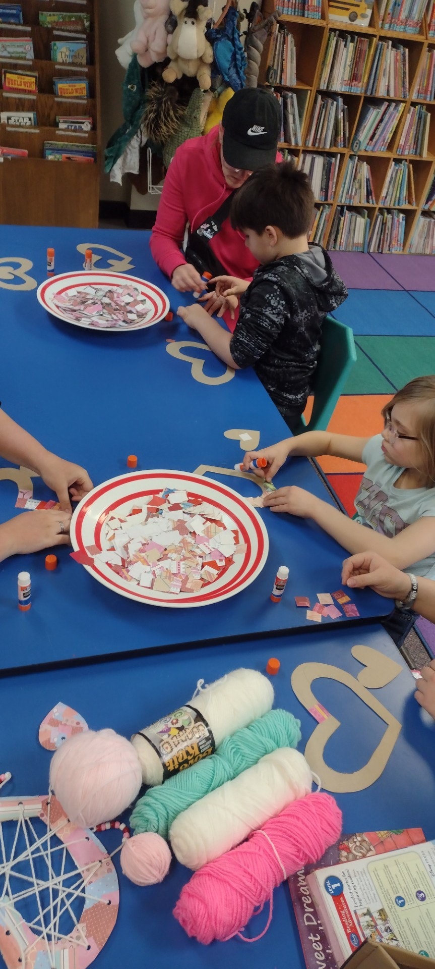 Kids and adults are crafting with paper, glue, and yarn at a colorful table filled with supplies in a library or activity space.