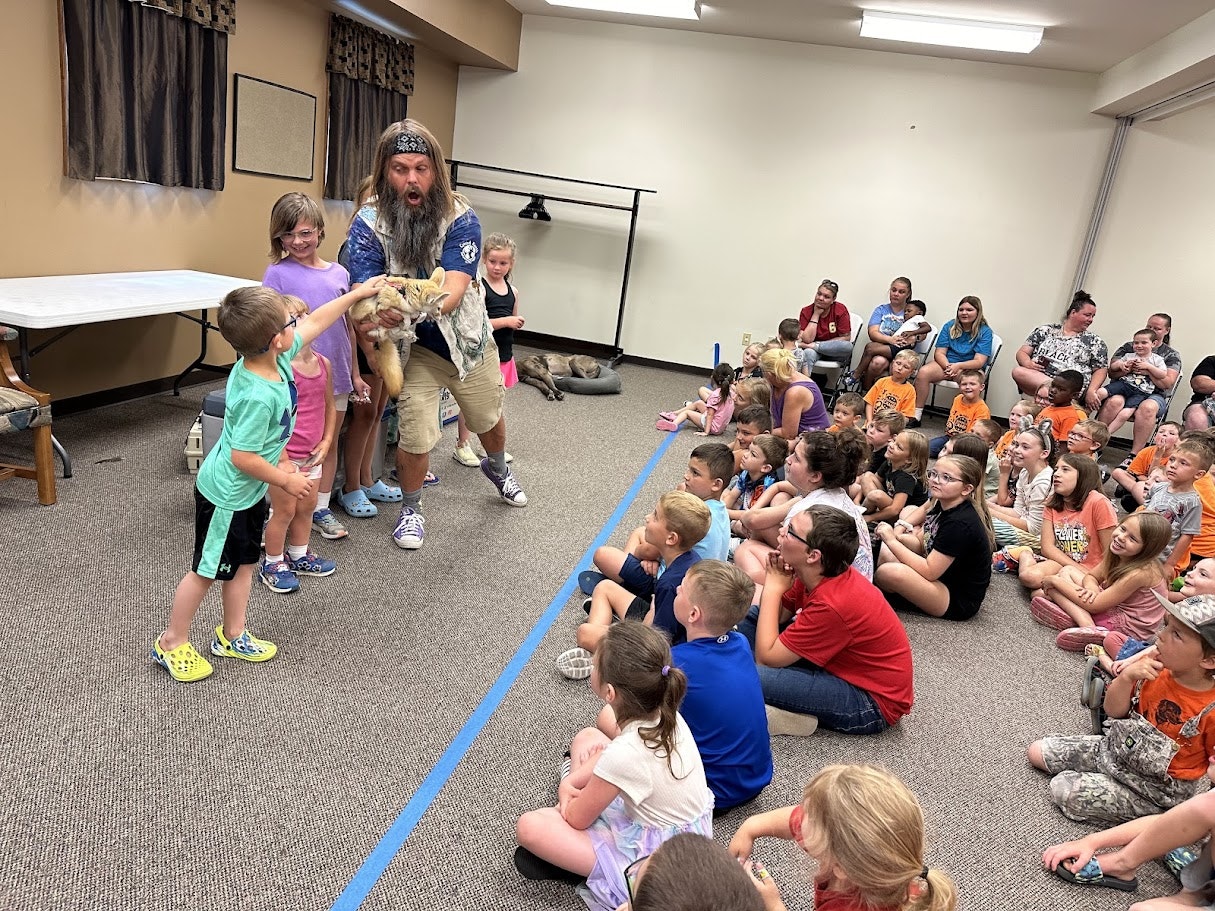 A man holds a small animal as children gather around to touch it, while seated kids and adults watch in a classroom setting.