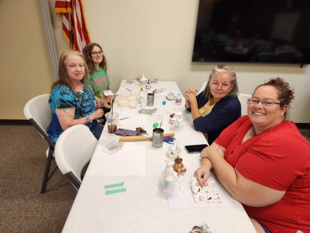 Four people sitting at a table with crafting supplies and beverages, smiling at the camera.