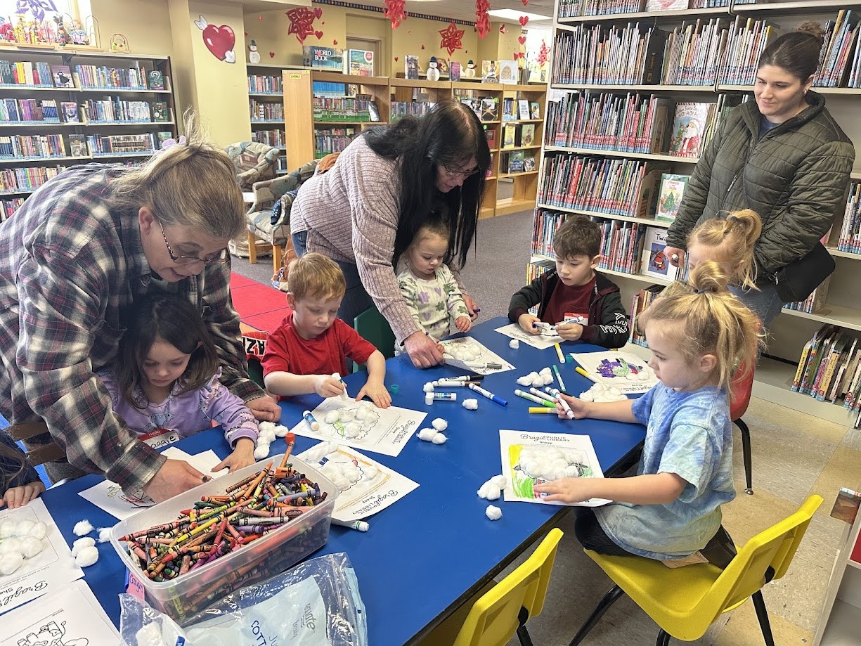 Children coloring with adults assisting in a library, with crayons and cotton balls on a table.