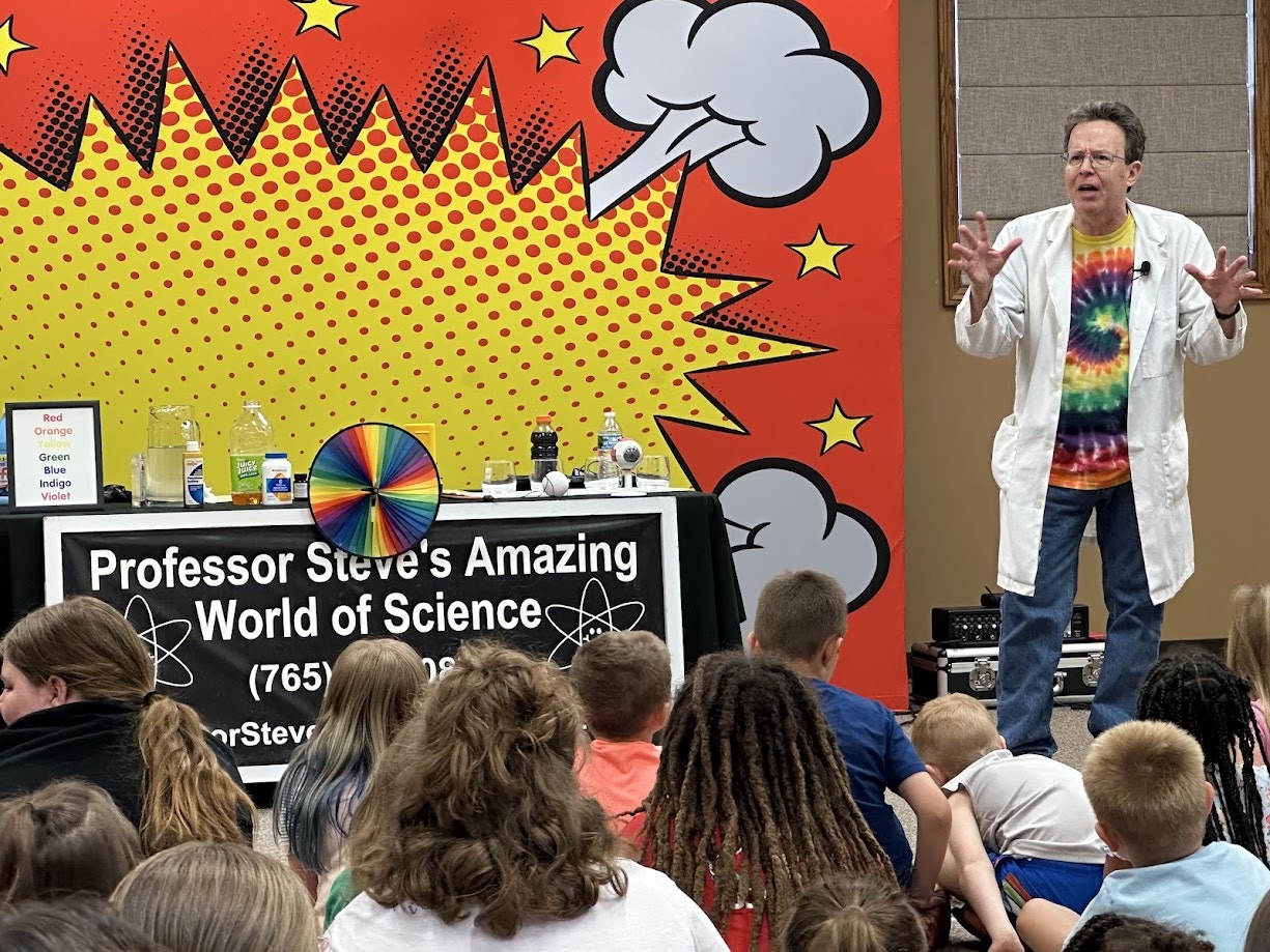 A man in a lab coat gives a science demonstration to children, with a colorful backdrop and table of experiments.