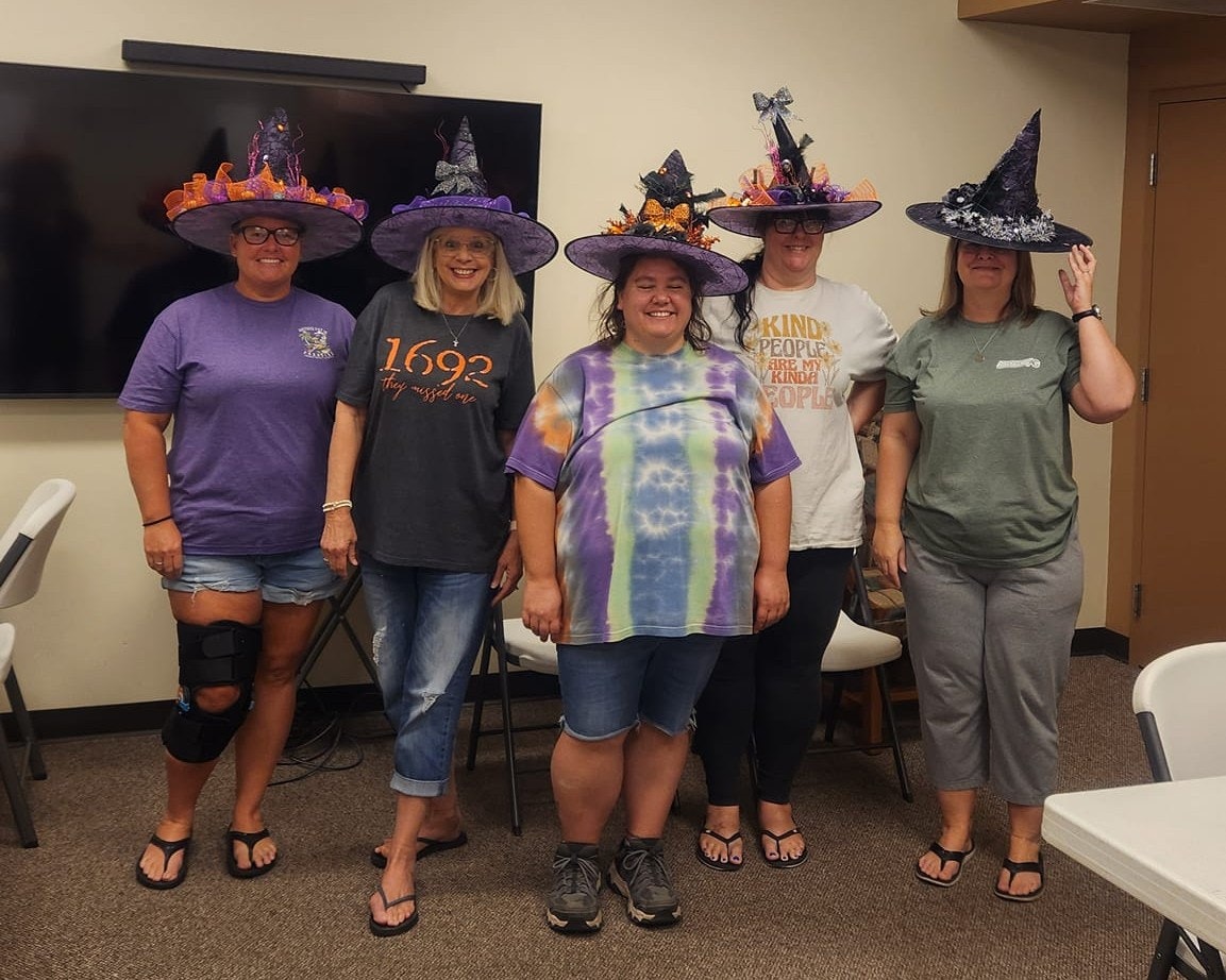 Five people wearing decorated witch hats in a room with chairs and a TV screen.
