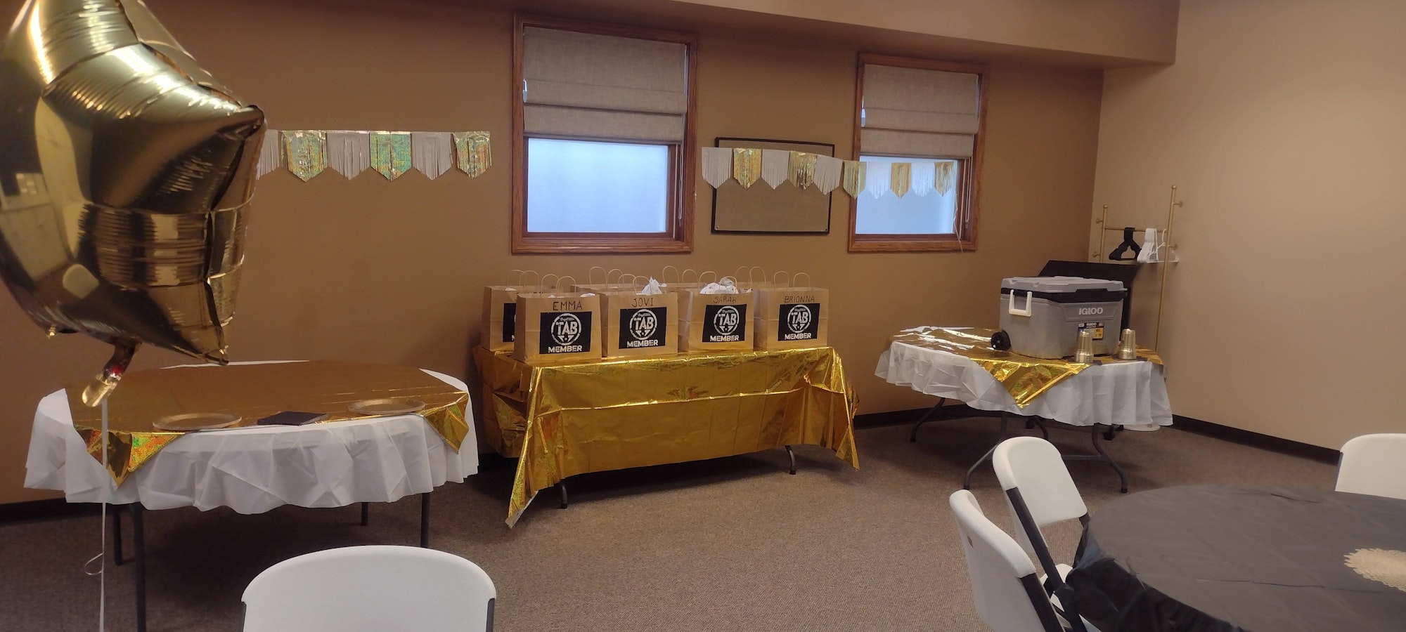Room set for a party with gold decorations, tables, gift bags labeled "Member," and a star-shaped balloon.