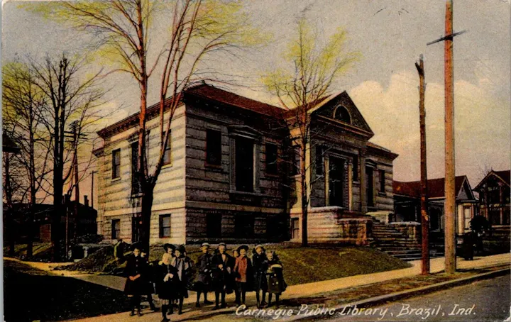 A group of people stands outside the Carnegie Public Library in Brazil, Indiana, surrounded by bare trees and a clear sky.