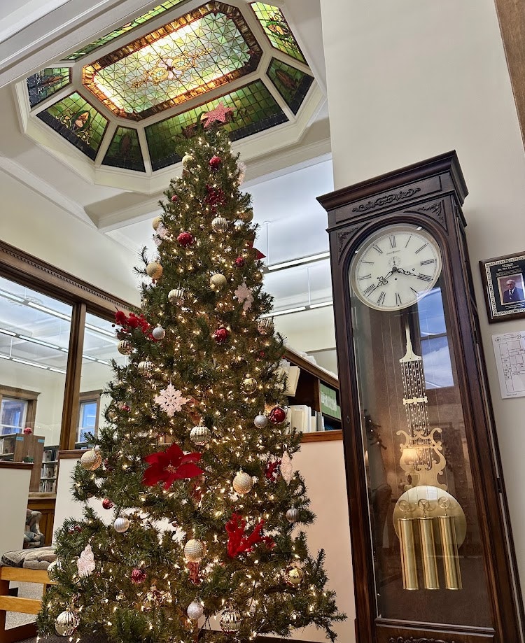 A decorated Christmas tree next to a tall wooden clock, under a stained glass ceiling.