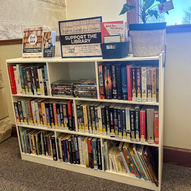 Bookshelf with various books and DVDs, "Support the Library" sign, plant on top, and donation box.