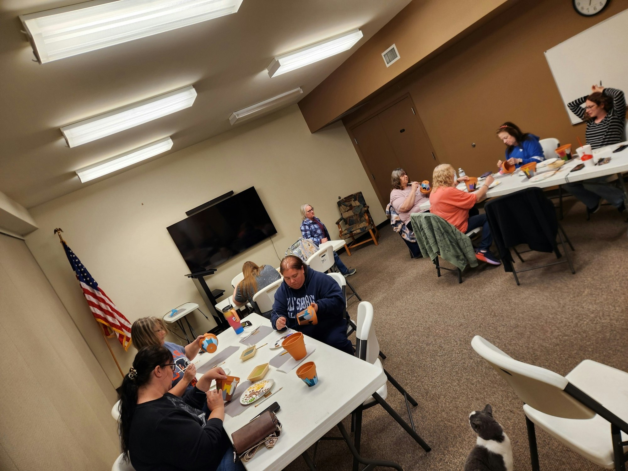 A group of people sitting around tables engaged in a craft activity, with a cat nearby and an American flag in the room.