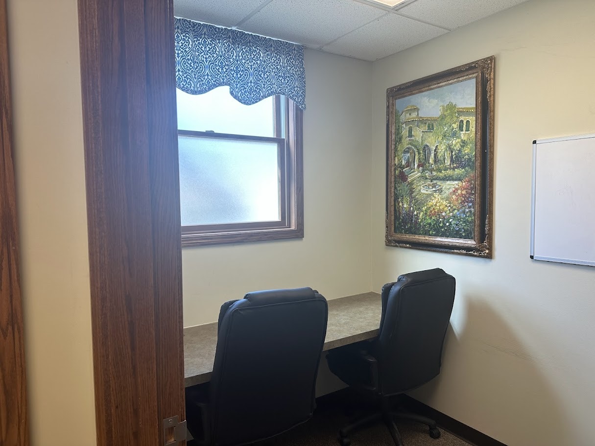 A small office with two chairs, a desk, a window with a curtain, a framed painting on the wall, and a whiteboard.