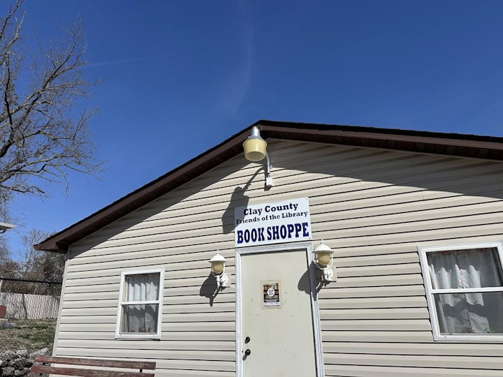 Small building with "Clay County Friends of the Library Book Shoppe" sign, two windows, and a door under a clear blue sky.