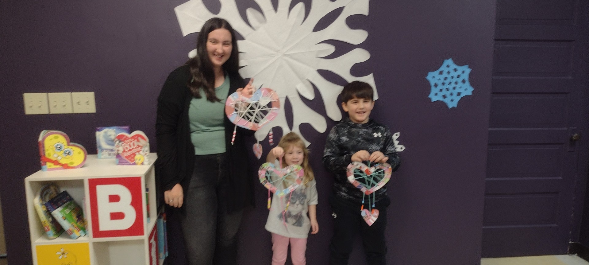 A woman and two children hold handmade heart crafts in front of colorful decorations on a purple wall. Fun, creative atmosphere!