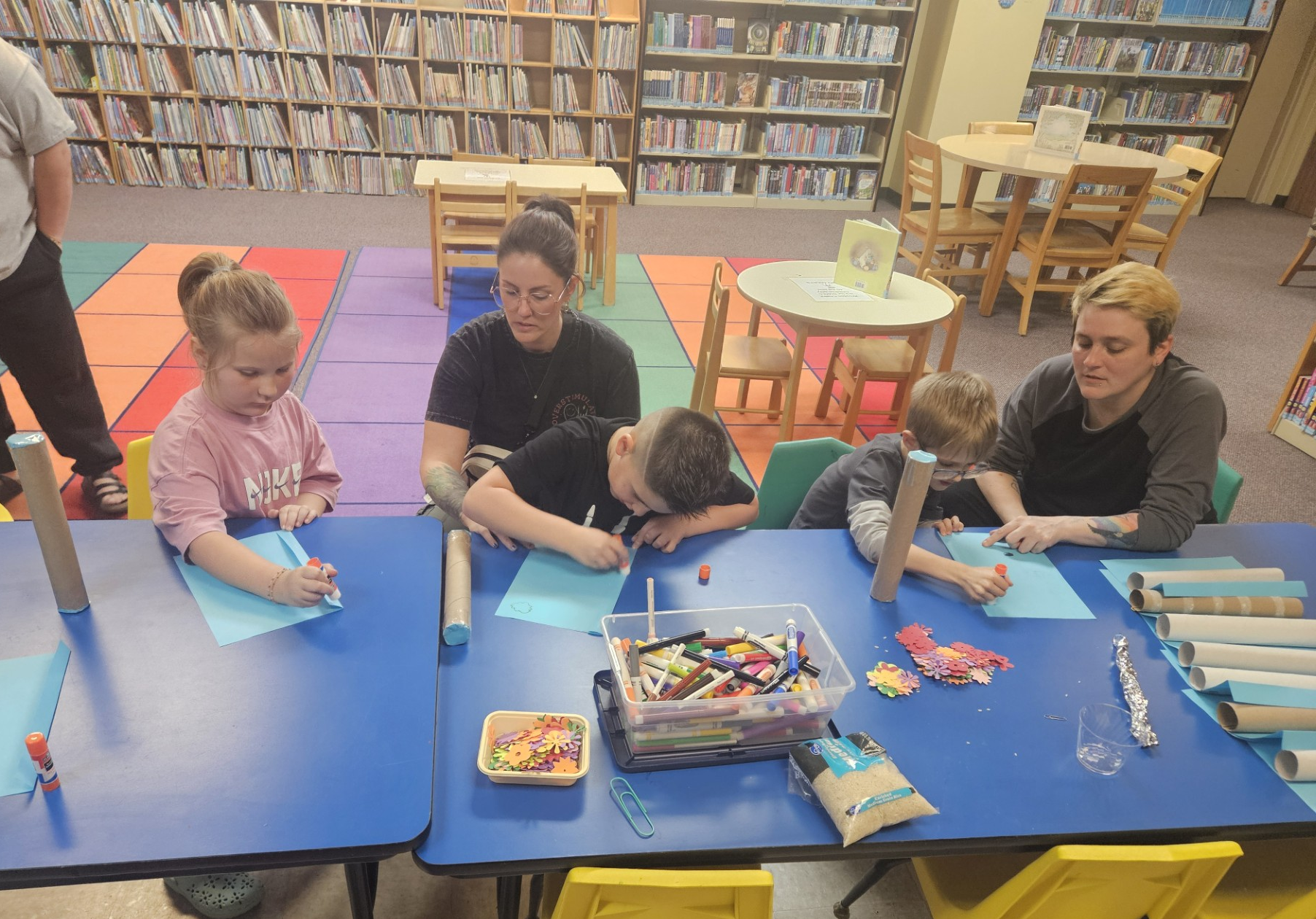 Children and adults are crafting with markers, paper, and supplies in a library setting. Brightly colored carpet and shelves of books are visible.