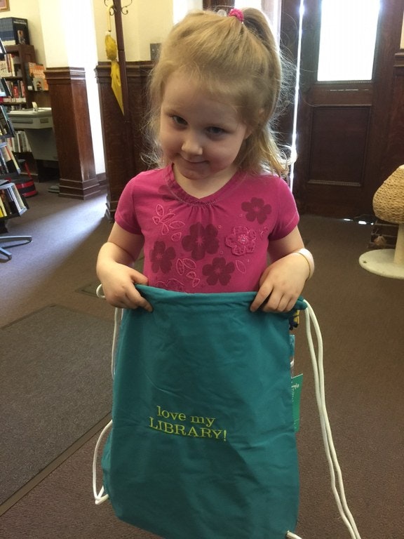 May contain: face, head, person, photography, portrait, clothing, t-shirt, bag, child, female, girl, book, indoors, library, and publication