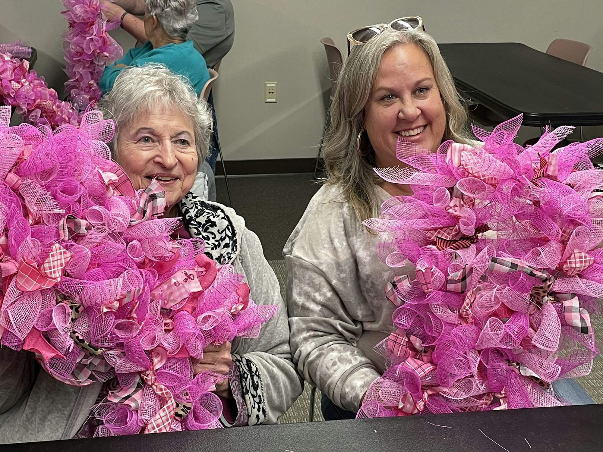 Two women are smiling while holding large, vibrant pink wreaths at a crafting event. Many other participants are in the background.