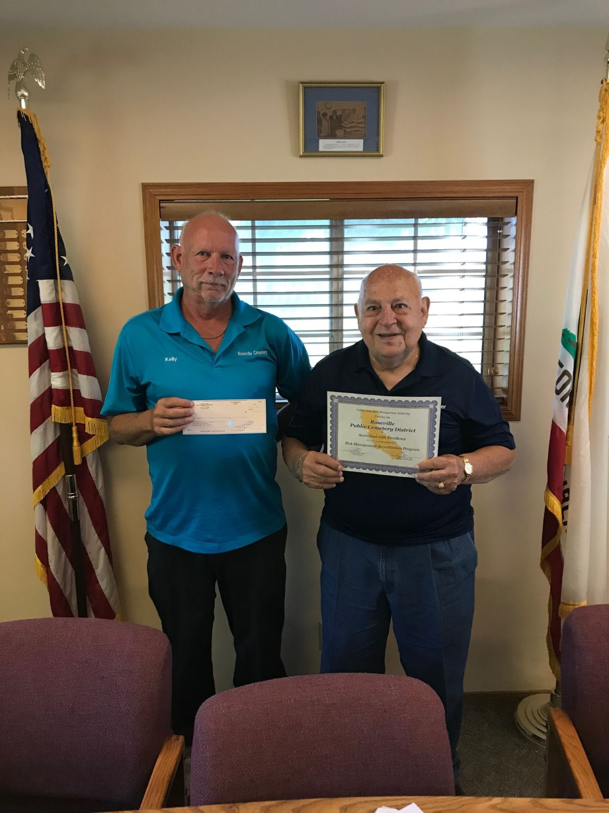 Two men standing indoors holding a check and a certificate, with flags and a framed picture in the background.