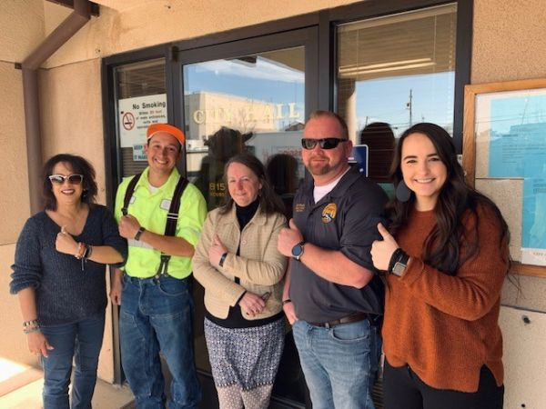 Five people standing in front of a glass door labeled "City Hall," all smiling and giving thumbs up.