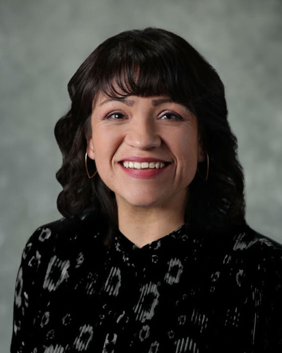 A person with dark hair is smiling, wearing a black patterned top and hoop earrings, against a neutral background.