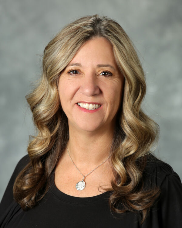 A person with long, wavy hair and a necklace, smiling against a gray background.