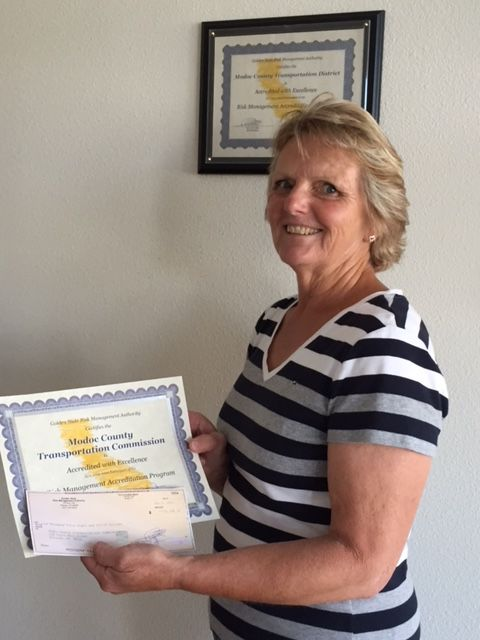 A woman holding a certificate from the Modoc County Transportation Commission, with another framed certificate on the wall.