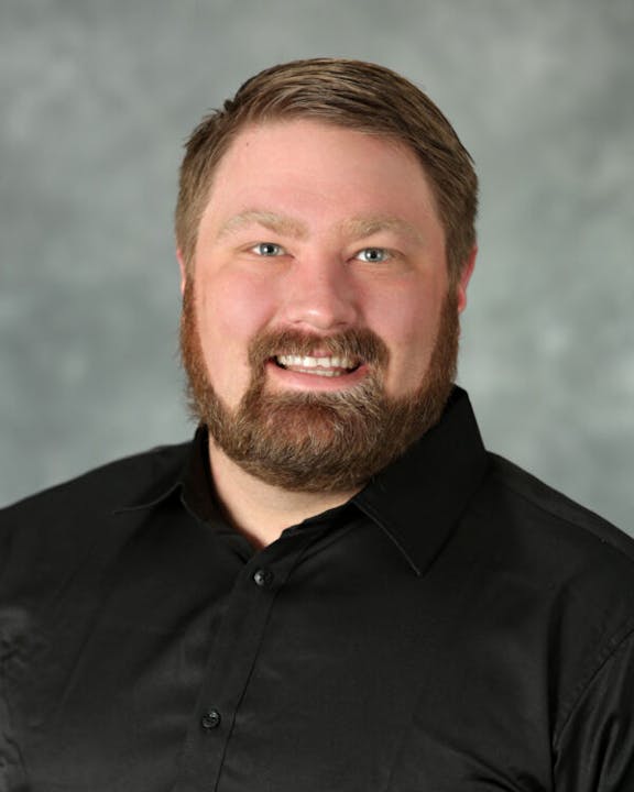 A man with a beard and short hair wearing a black shirt, smiling against a gray background.