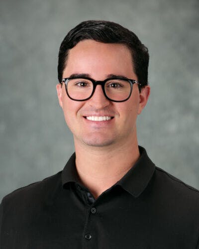 A person with dark hair and glasses, wearing a dark polo shirt, smiling against a neutral background.