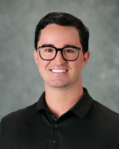 A person with dark hair and glasses, wearing a dark polo shirt, smiling against a neutral background.