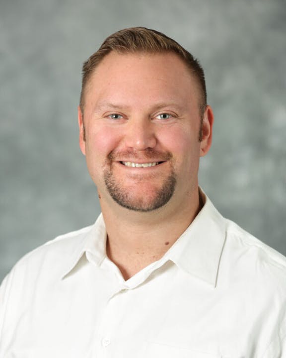 A person smiling, wearing a white shirt against a gray background.