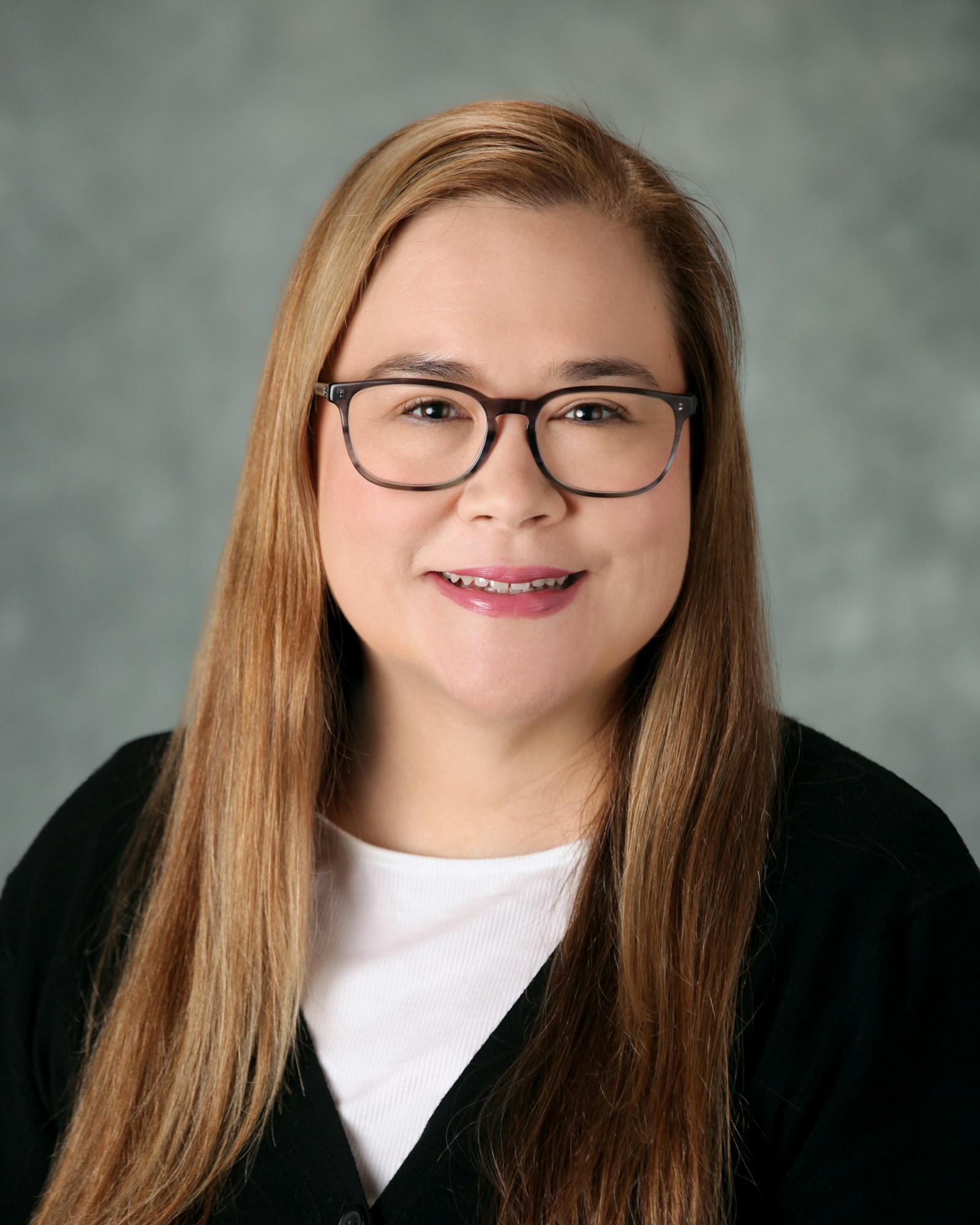 The image shows a woman with long hair, wearing glasses and a black jacket, smiling against a soft gray background.