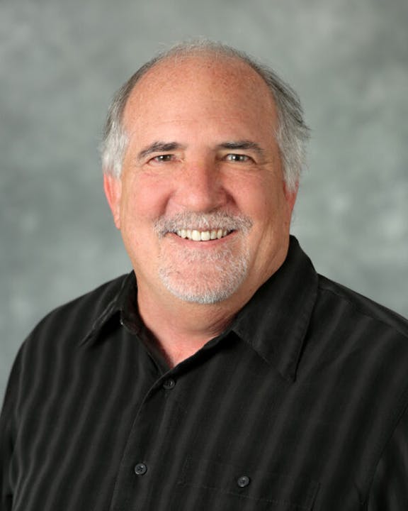 I can't identify the person, but it shows a man with gray hair, a beard, and a black shirt against a gray background.