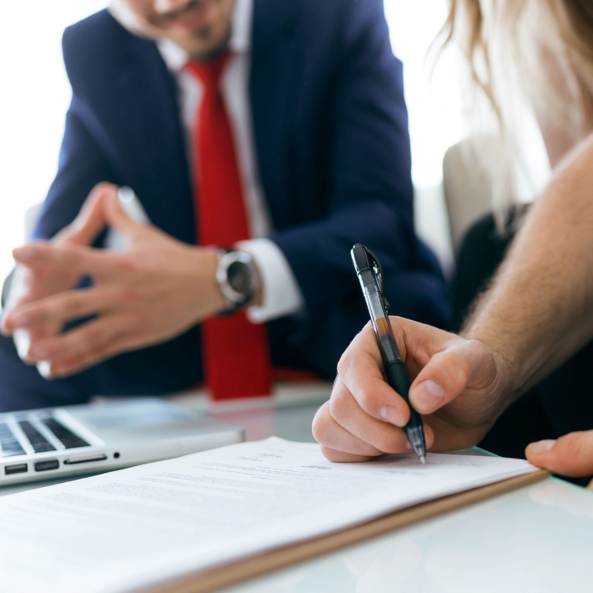 Two people in professional attire, one signs a document, while the other observes near a laptop.
