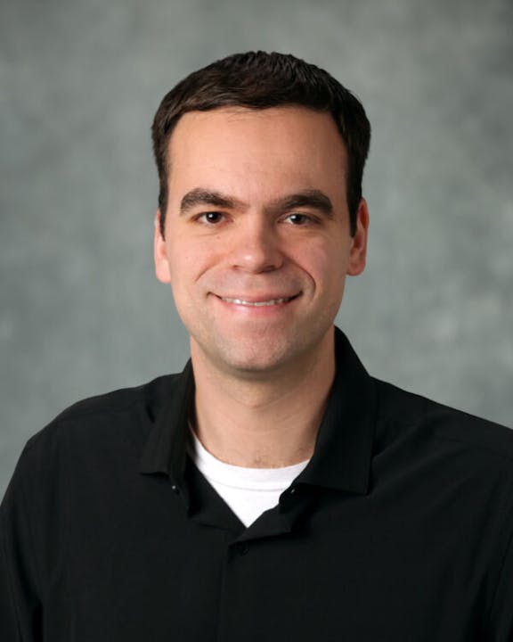 A person smiling, wearing a black shirt and white undershirt, with a neutral background.