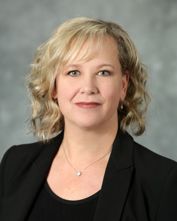A person with blonde hair, wearing a black blazer and a necklace, poses against a neutral background.