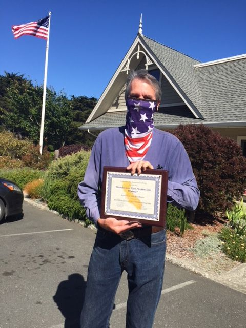 A person wearing a flag-patterned face covering holds a framed certificate outdoors near a house and flagpole.