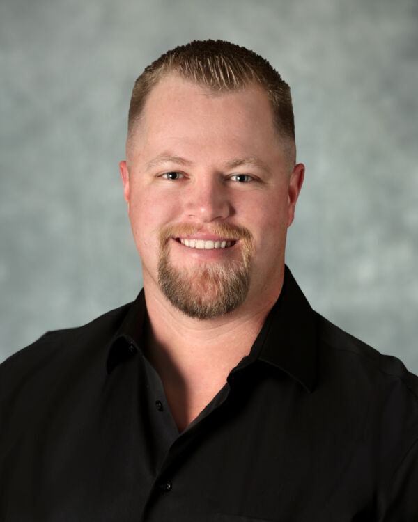 A person with short hair and a goatee, wearing a black shirt, smiling against a gray background.